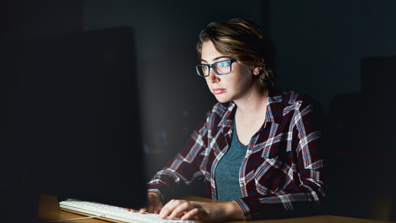 A young woman working by the light of her computer monitor looks serious