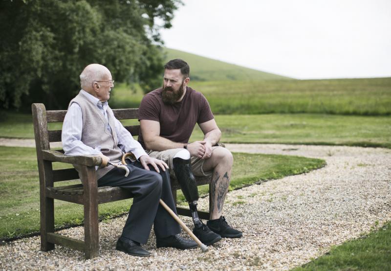 Dans le cadre du programme Blesma, un jeune vétéran portant une prothèse à la jambe est assis sur un banc de parc et discute avec un vétéran âgé portant une prothèse au bras