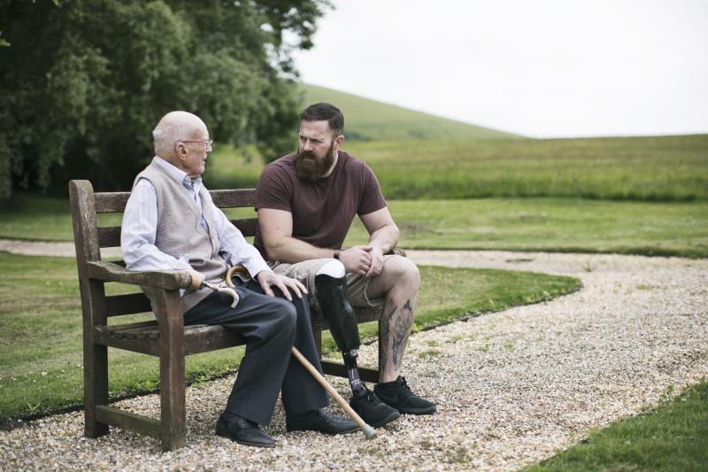 A young veteran with a prosthetic leg sits on a park bench talking to an elderly veteran with a prosthetic arm, as part of a Blesma program that offers help to veterans